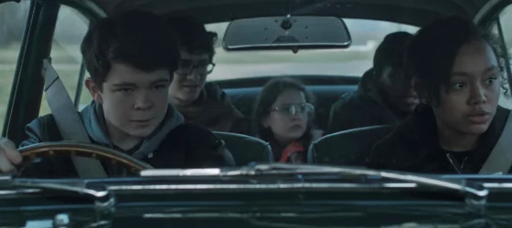 Monty driving a car next to Jamila with Darwin, Penny, and Alfie sat down in the back.